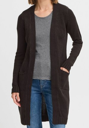 Mujer con un cárdigan largo de punto negro sobre una camiseta gris y jeans azules, con una mano en el bolsillo del cárdigan.