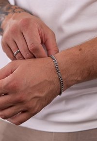 Silberne Metallkette als Armband am Handgelenk, mit ineinandergreifenden Gliedern und glänzender Oberfläche. Hauttextur und ein Ring an der anderen Hand sichtbar.