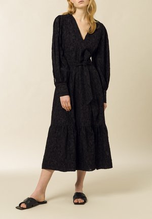 Robe noire à manches longues avec un col en V, en tissu texturé, taille cintrée avec un lien, et jupe à volants. Associée à des sandales noires plates.