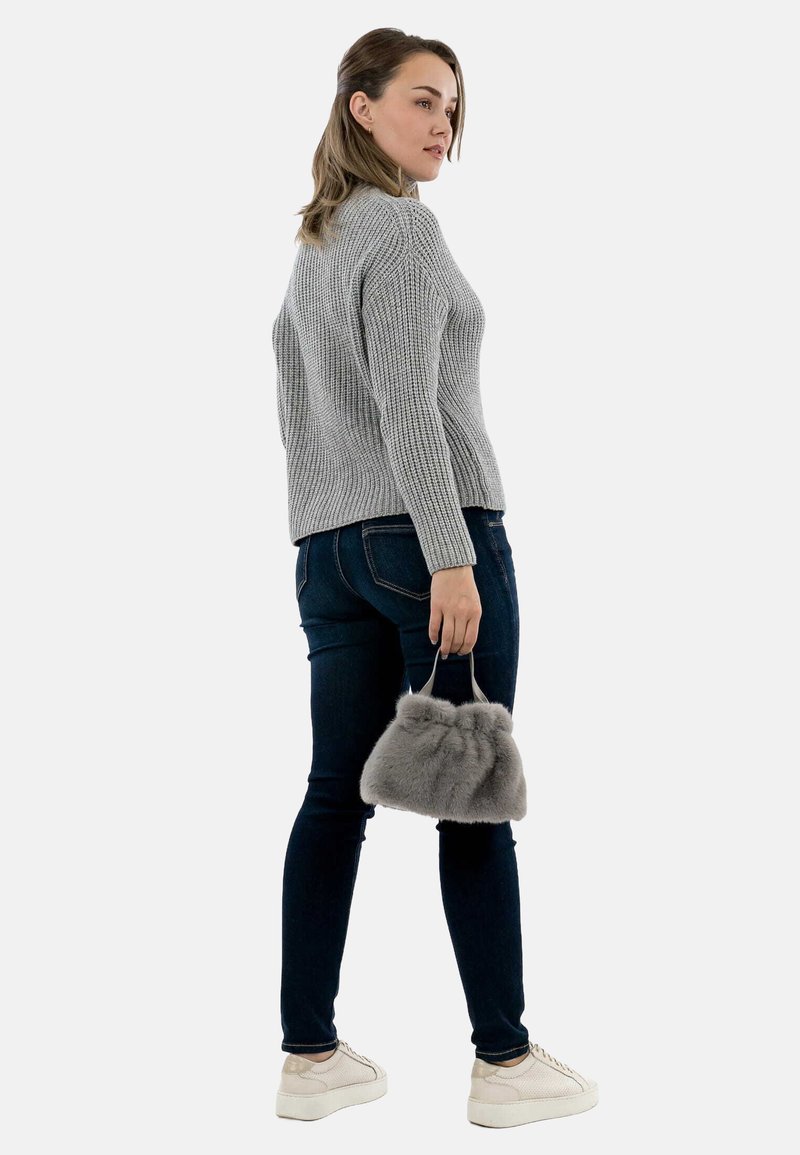 Donna che indossa un maglione grigio a maglia e jeans scuri, tiene una borsa in pelliccia grigia, in piedi e guarda oltre la spalla.