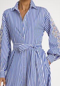 Vestido camisa de algodão às riscas azuis e brancas, com fecho de botões, cinto de atar na cintura e detalhes em renda nas mangas compridas.