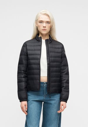 Blondína v čiernej prešívanej bunde cez biely krátky top a modré džínsy, stojaca pred jednoduchým svetlým pozadím.