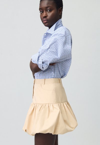 Une chemise à carreaux bleus est portée avec une jupe évasée de couleur crème. La jupe présente un ourlet froncé et une poche avant.