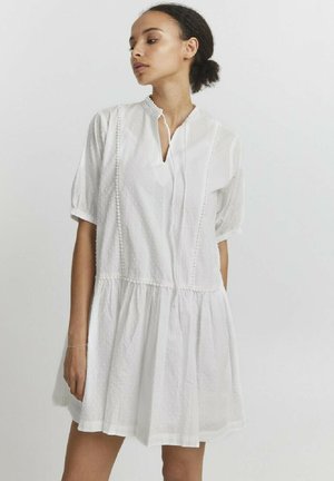 Femme aux cheveux attachés portant une robe blanche texturée à manches courtes avec une jupe froncée et un décolleté fendu, se tenant devant un arrière-plan uni.