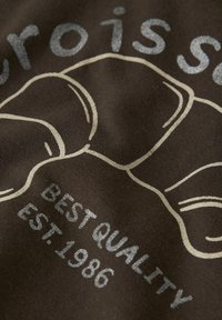 Brązowy materiał z graficznym wzorem na cieszące się popularnością rogaliki w jasnobeżowym konturze, z napisem: "NAJLEPSZA JAKOŚĆ EST. 1986" w srebrnym kolorze.