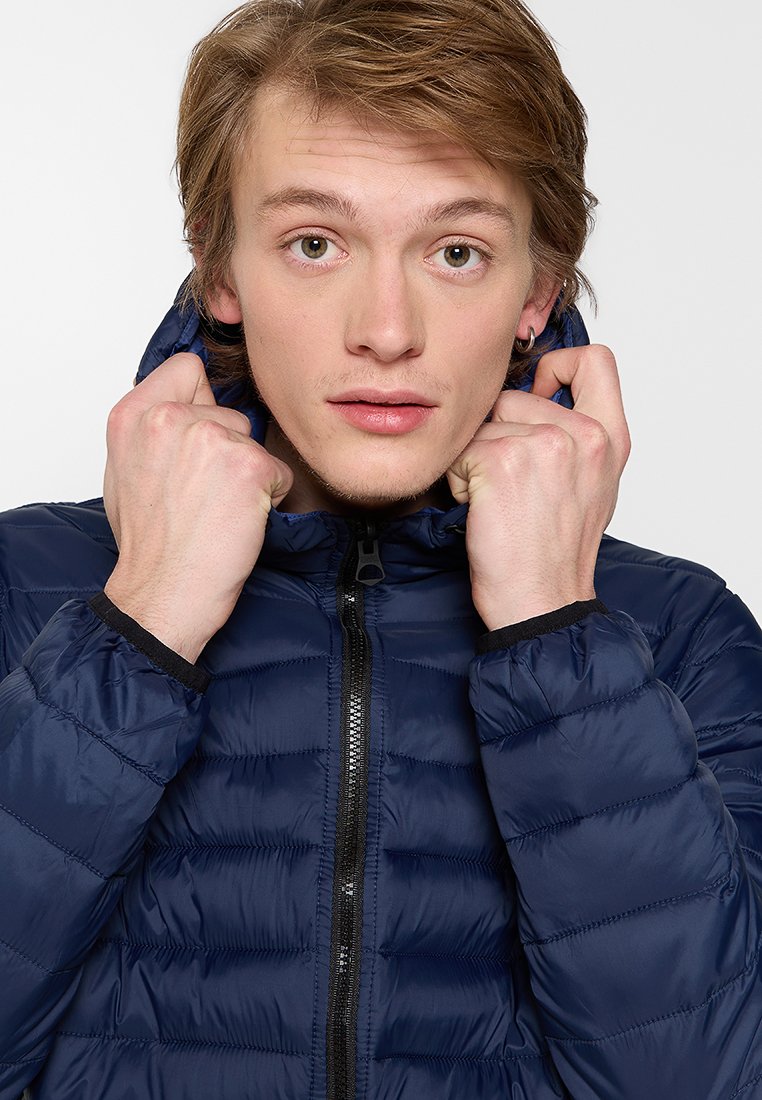 Chaqueta acolchada azul marino con cremallera frontal, puños elásticos y capucha, que presenta un sutil diseño acolchado y una textura suave.