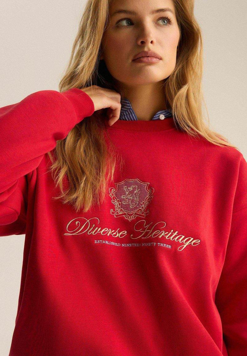 Czerwony sweter z długim rękawem, żebrowanym kołnierzem i haftowanym logo "Różnorodne Dziedzictwo" w kolorze złotym. Nałożony na niebieską koszulę w paski.