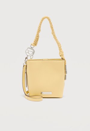 Sac à main en forme de seau jaune avec une poignée en corde torsadée, une bandoulière amovible et un charm argenté en forme de visage portant des lunettes de soleil.