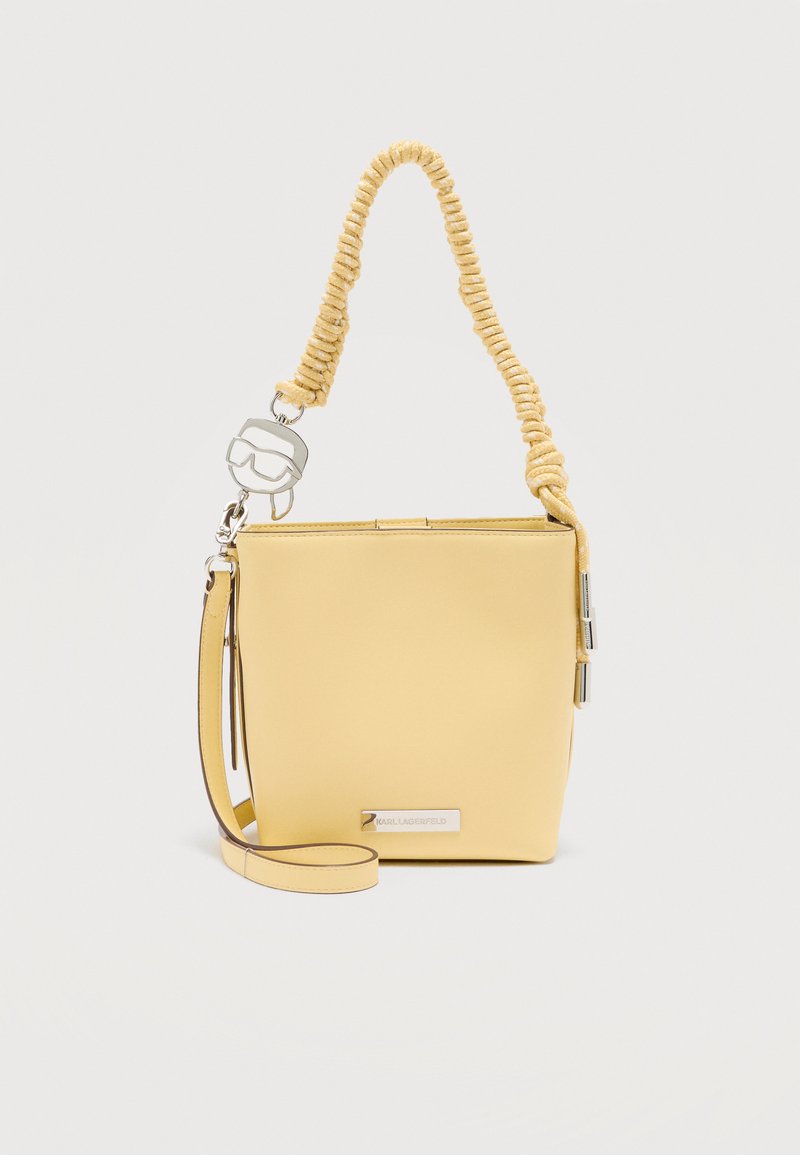 Κίτρινη τσάντα χειρός σε σχήμα κάδου με στριφτή λαβή σχοινιού, αποσπώμενο ιμάντα ώμου και ασημένιο γούρι σε σχήμα προσώπου που φορά γυαλιά ηλίου.