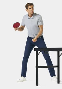 Grijze polo, marineblauwe broek en witte sneakers. Houdt een rode pingpongbat en een oranje bal vast naast een zwarte pingpongtafel.