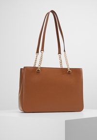 Bruine leren tote bag met geperforeerde textuur, dubbele slanke schouderbanden en gouden kettingaccenten voor extra detail.