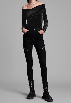 Woman wearing an off-shoulder black velvet top, high-waisted black pants with zip details, and black lace-up ankle boots, standing with hands in pockets.