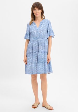 Woman standing wearing a blue patterned knee-length dress with flutter sleeves and beige flat sandals on a plain background.