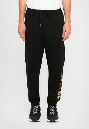 Man wearing black jogger pants with gold "COUTURE" text on leg and black and white branded sneakers.