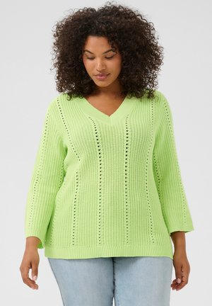 Lichtgroene gebreide trui met een V-hals, voorzien van een open steekpatroon en driekwart mouwen, gecombineerd met lichtblauwe jeans.