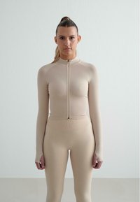 Beige long-sleeve top with a high collar, half-zipper, and ribbed texture; paired with matching high-waisted leggings.