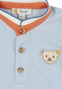 Lichtblauw polo shirt met een kraag met oranje accenten, twee knopen en een geborduurd berengezicht logo op de borst. Structuurstof.