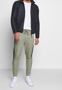 Schwarze gesteppte Jacke über einem weißen strukturierten T-Shirt, kombiniert mit hellgrünen Jogginghosen mit schwarzen Seitenstreifen und weißen Sneakers.