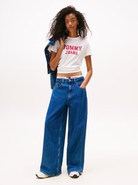 Weißes T-Shirt mit kurzen Ärmeln und rotem "TOMMY JEANS"-Logo, kombiniert mit lockeren blauen Jeans und weißen Sneakern. Glatte Textur, lässige Passform.