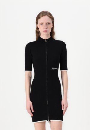 Schwarzes geripptes Kleid mit einem Reißverschluss vorne, kurzen Ärmeln und weißen Akzenten an den Ärmelbündchen und dem Saum. Mit einem Logo auf der Brust.