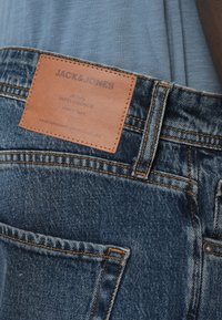 Τζιν denim με υφή μπλε υφάσματος, με πορτοκαλί ραφές, δερμάτινη ετικέτα με ανάγλυφο το "JACK & JONES" και πίσω τσέπη.