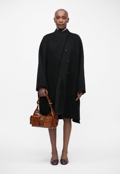 Casaco de lã preto com gola alta e mangas oversized; mala de couro castanho com múltiplos bolsos; sapatos escuros.