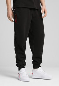 Czarne dresowe spodnie o slim fit, wykonane z miękkiego materiału, z czerwonym logo "Formula 1" z boku i elastycznymi mankietami. Noszone z białymi tenisówkami.
