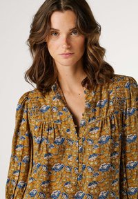 Bluse in Senfgelb mit blauem Blumenmuster, mit V-Ausschnitt, Knopfleiste und strukturiertem Stoff. Lange, aufgeblähte Ärmel.