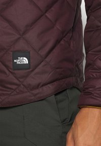 Bordová prošívaná bunda s černou nášivkou loga a bílým textem. Hladký, lesklý materiál s zaobleným lemem a podrobnostmi v ribbovém designu na límci.