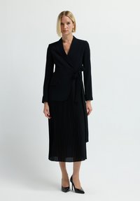 Frau mit glattem blonden Haar, die ein schwarzes Wickelblazer, einen plissierten Midirock und schwarze High Heels trägt, steht vor einem schlichten Hintergrund.