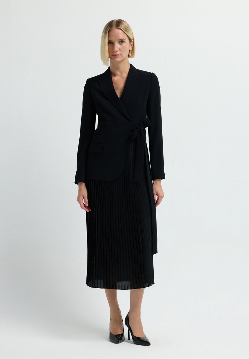 Frau mit glattem blonden Haar, die ein schwarzes Wickelblazer, einen plissierten Midirock und schwarze High Heels trägt, steht vor einem schlichten Hintergrund.