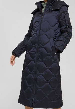 Langer, gesteppter schwarzer Wintermantel mit wellenförmiger Steppung, Frontreißverschluss, Seitentaschen und hohem Kragen, getragen über einem hellgrünen Rollkragenpullover.