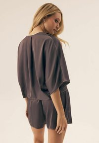 Kobieta z długimi blond włosami, ubrana w luźny, ciemnoszary top z krótkim rękawem i pasujące szorty, stoi i patrzy przez ramię.