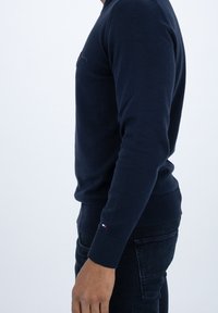 Granatowy sweter dziany z okrągłym dekoltem, ściągaczami na mankietach i dole. Mały detal logo na piersi. Miękka faktura i dopasowany krój.