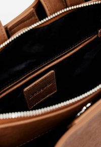 Interno di una borsa a mano in pelle marrone con foderato nero, chiusura con zip e un piccolo etichetta con il marchio. Presenta superfici lisce e testurizzate.