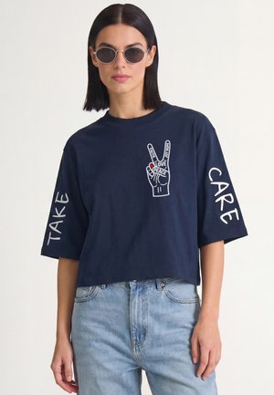 Femme portant des lunettes de soleil rondes et une chemise bleu marine avec un motif de main de paix et les mots "Take Care" sur les manches.