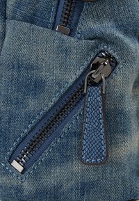 Tela vaquera en azul claro con textura suave, que presenta una cremallera negra y una lengüeta decorativa de cuero con un patrón texturizado.