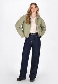 Giacca bomber verde oliva trapuntata con un design corto, indossata sopra un maglione crema e abbinata a jeans blu scuro a vita alta e stivali neri.