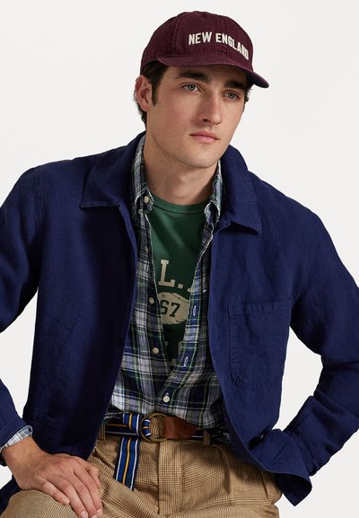 Navy linen jacket over a plaid button-up shirt, green graphic t-shirt, beige checkered pants, and a maroon cap with white text.
