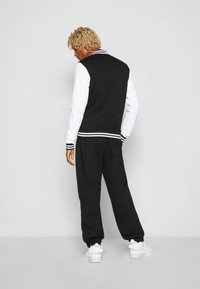 Chaqueta tipo varsity en blanco y negro con un tejido suave, puños rayados y joggers negros a juego. El modelo lleva zapatillas blancas.