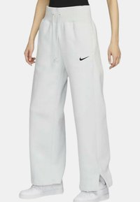 Bílé tepláky Nike s elastickým pasem na šňůrku, bočními kapsami a černým logem na levé nohavici. Mají volný střih a otevřené lemy.