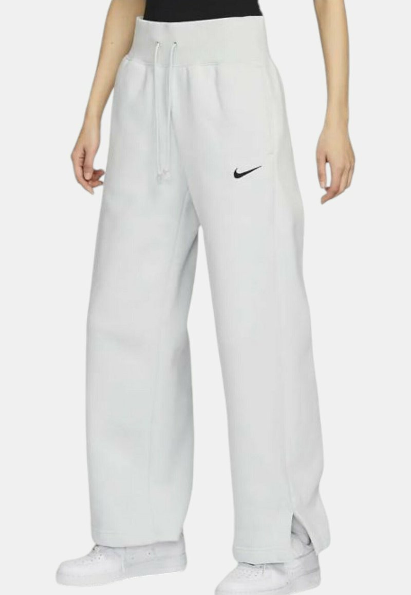Bílé tepláky Nike s elastickým pasem na šňůrku, bočními kapsami a černým logem na levé nohavici. Mají volný střih a otevřené lemy.