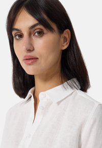 Camicia bianca in lino con colletto classico e chiusura a bottoni. Il tessuto ha un aspetto morbido e texturizzato con sottili dettagli di tessitura.