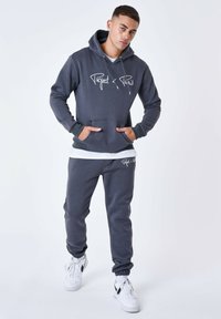 Sudadera gris con capucha, con bolsillo frontal y logo blanco. Combinada con pantalones de chándal a juego. Modelo lleva zapatillas blancas.