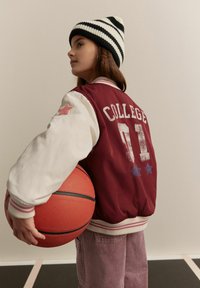 Burgundi varsity jope, valgete varrukatega, "COLLEGE 01" print, roosa tähe detailiga, koos punase korvpalliga ja ribadega beanie'ga.