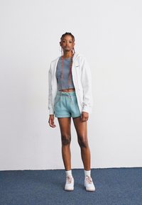 Lys grå hettejakke med glidelås, tealkorte bukser og grå crop topp. Hvite sneakers med rosa detaljer. Lang flette, står på blått teppe.