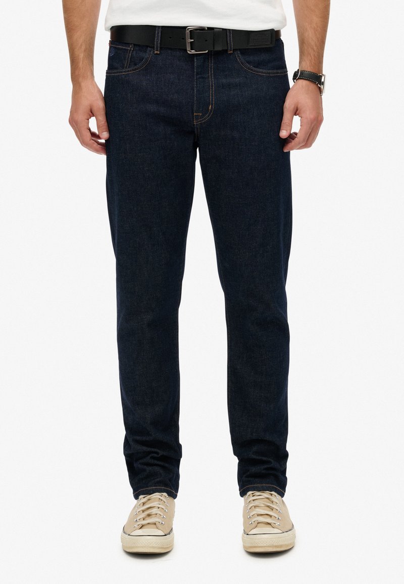 Jeans de mezclilla azul oscuro con un corte recto, costuras naranjas, bolsillos frontales y un cinturón negro. Zapatillas blancas con cordones beige completan el look.