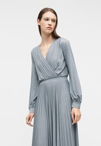 Γυναίκα που φοράει μακρυμάνικο, μεταλλιζέ ασημί πλισέ φόρεμα με ζωνάρι και εφαρμοστά μανσέτα, στέκεται μπροστά σε απλό λευκό φόντο, κοιτάζοντας αριστερά.