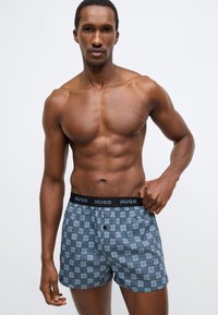 Μπόξερ σε σκούρο γκρι με σχέδιο πλέγματος που περιλαμβάνει το όνομα της μάρκας. Ελαστική ζώνη μέσης με την ένδειξη "HUGO" σε μαύρο χρώμα. Υλικό από βαμβάκι.