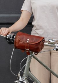 Borsa da bicicletta in pelle marrone con pattina, hardware argentato e fibbia. Attaccata al manubrio della bicicletta, con impugnatura e leve dei freni visibili.